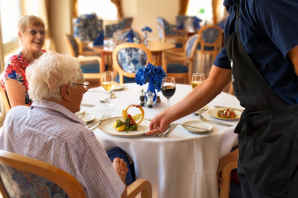 A server presents an elegant meal to a smiling elderly man at a dining table, with two women and drinks in the background.